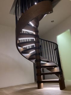 Dark oak wood-wrapped spiral stair with black metal railing and LED strips under each tread.