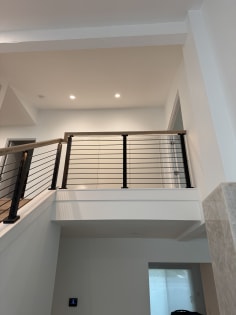 Black horizontal rod railing with a light oak handrail lines an interior balcony and stair in a bright white home.