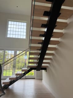 View under a black mono stringer carrying white-oak treads with glass railing in front of tall grid windows.