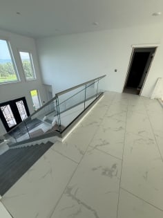 Frameless glass with a gray top cap rings a stair opening on a white marble-tile floor below tall windows.