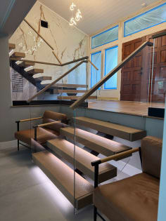 Thick oak floating treads lit from below with frameless glass railing and wood handrail beside leather lounge chairs.