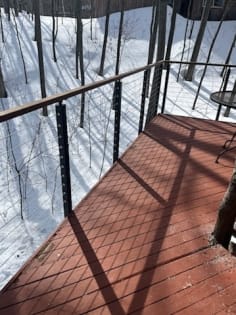 Side-mounted cable railing with wood handrail edges a red deck above a snowy wooded slope.