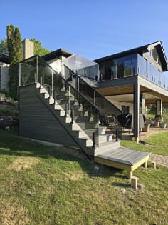 Glass panels on black talon clamps follow an exterior deck stair and wrap an upper deck on a lakeside home.