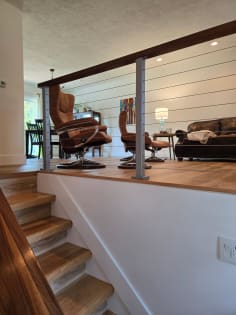 Silver-post cable railing with a wood cap rail edges a raised living area above an oak staircase.