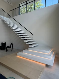White floating treads rise from stacked LED-lit plinths beside frameless glass and a black handrail in a tall gallery.