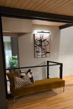 Black post-to-post glass railing guards a stair opening beside a velvet bench on hardwood floors.