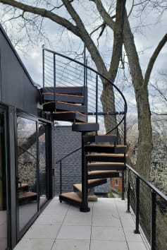 Black exterior spiral staircase with wood-look treads rising from a tiled balcony lined with black cable railing among trees.