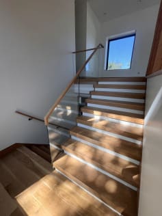 Dappled sunlight falls across a white oak switchback stair with a frameless glass guard, wood handrail and wall handrail.