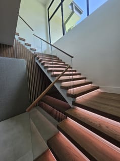 Wood treads with glowing LED strips and a frameless glass railing with wood handrail climbing beneath a tall window.