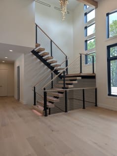 White oak floating treads on a black mono stringer switch back at a landing, with LED under-lighting and black cable railing below tall windows.