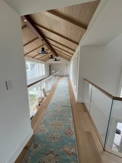 Frameless glass railing with a white oak cap rail lines a long vaulted upstairs hallway with a runner rug.