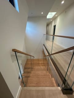 View down an open wood stair lined with frameless glass railing and white oak handrails under skylights.