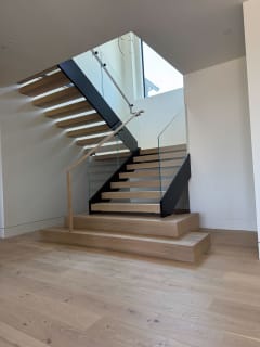 Light oak floating treads on a black mono stringer turn at a landing above a wood plinth, with glass railing and a wood handrail.