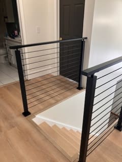 Black rod railing wraps the top of a stair opening on a light wood floor in a hallway.