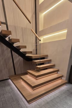 Floating light oak stair with frameless glass guard rising from stacked plinth boxes underlit with LED on stone tile.