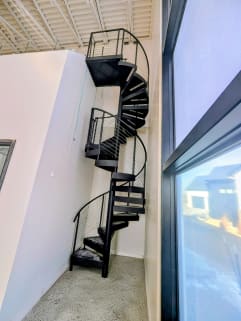 A black steel spiral stair with cable infill climbs beside a tall window in a white interior.