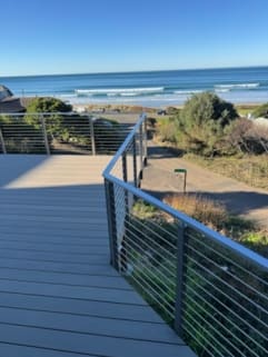 Brushed stainless posts and top rail with horizontal cable infill line a composite deck walkway overlooking the ocean surf.