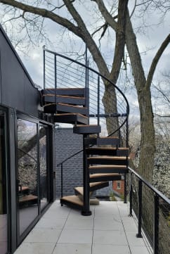 Black exterior spiral staircase with wood-look treads rising from a tiled balcony lined with black cable railing among trees.
