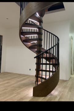 Spiral stair with a wood ribbon stringer, LED-lit treads and black vertical square balusters in a white room.
