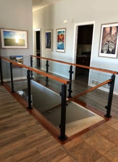 Black posts with glass panels and a stained wood top rail surround a stair opening in a hallway with framed art.