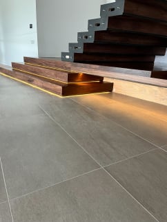 Stacked walnut plinth steps with warm LED under-glow at the foot of a cantilevered walnut stair on tile floor.