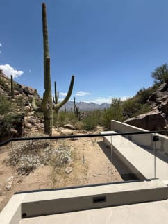Frameless glass with a black top rail guards a concrete terrace among saguaro cacti and rocky hills.