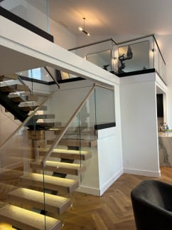 Light oak floating treads with glowing under-tread LEDs climb beside frameless glass with a wood cap rail to a glass-railed loft.
