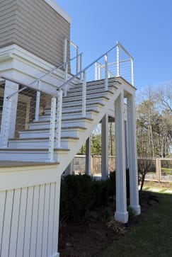 Exterior staircase with white posts and cable railing climbing to a second-story deck on a shingled home