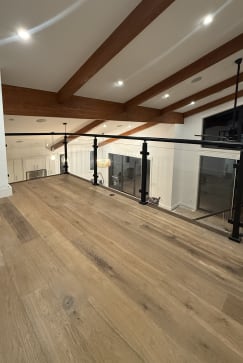 A black post-to-post glass railing lines an open loft with wide-plank oak floors and exposed beams.