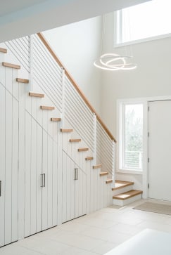 A white board-and-batten stair with oak treads and slim white cable railing beneath a ring pendant light.