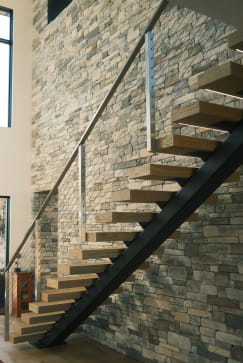 Light oak treads on a black mono stringer with stainless cable railing climb a stacked-stone wall.