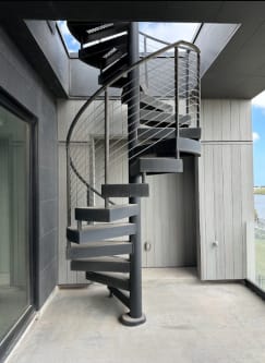 Dark exterior spiral staircase with stainless cable railing rising to a roof opening in a covered patio.