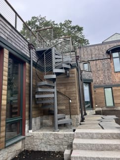 Gray steel spiral stair with cable railing climbs to a roof deck beside wood siding and granite steps.