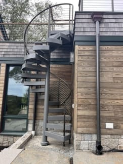 Gray steel spiral stair with curved cable railing beside cedar shingle and wood-clad walls.