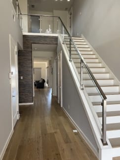 Stainless posts with horizontal rod infill and a matching handrail run up a white straight stair beside a long wood-floored hallway.