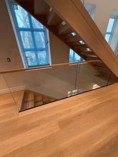 Frameless glass and oak handrail wrap a stair opening below a blue-taped arched window