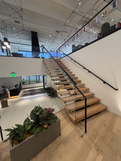 Wide wood floating treads with frameless glass and black handrails rising through a bright office lobby