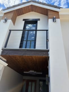A black cable railing Juliet balcony on a white stucco facade beneath a wood-clad gable.