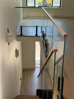View down an oak stair lined with glass on black talons and a wood handrail to a talon-glass landing guard.