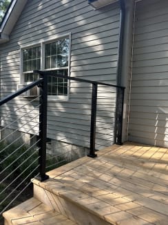 Black cable railing wraps a small cedar entry landing against gray lap siding.