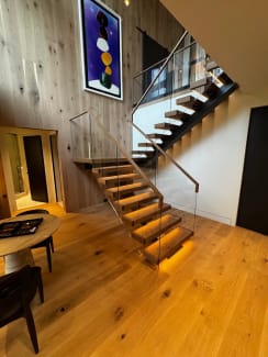 Thick oak treads with warm under-tread LED lighting float on black stringers beside frameless glass with a wood handrail in a double-height wood-paneled room.
