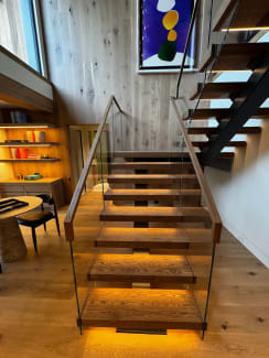 A straight run of oak floating treads with LED under-lighting rises between frameless glass guards topped with wood handrails.