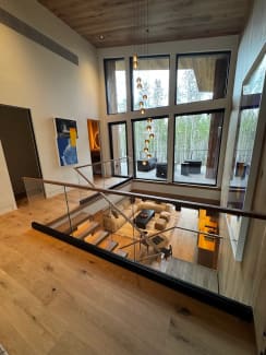 A frameless glass guard with wood cap overlooks a double-height living room with floor-to-ceiling windows.