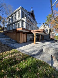 A raised wood deck with black posts and glass infill sits above a stone patio at a gray-sided lake house.