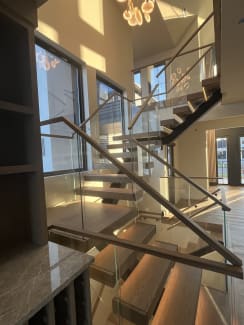 Oak floating treads on a dark stringer climb between frameless glass panels with a wood handrail in warm evening light.
