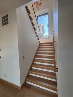 A white oak stair with white risers rises toward a tall window between a wall-mounted wood handrail and a frameless glass guard.
