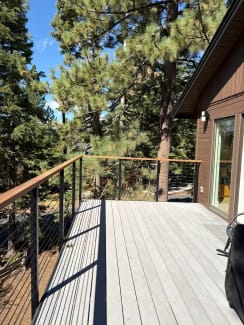 Wood-capped black cable railing wraps a gray deck among tall pines beside a brown-sided cabin.