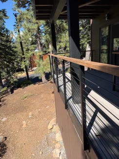 Wood-capped cable railing runs along an elevated deck on a dark-sided cabin among pines.