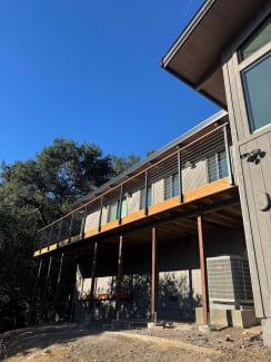 A long elevated wood deck with black posts, wood cap and cable infill runs along a gray home under oak trees.