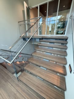 Floating dark wood treads with frameless glass and stainless handrails climb toward a glass curtain wall.